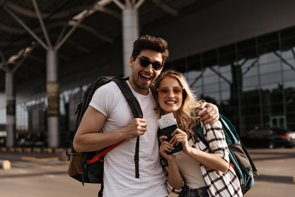young brunette man smiles widely hugs his attractive girlfriend couple travelers holds backpacks passports airport background